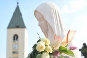 Statua Madonna Medjugorje con fiori in mano e chiesa sullo sfondo